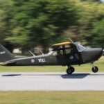 A military aircraft in motion as it lands on a runway surrounded by greenery.